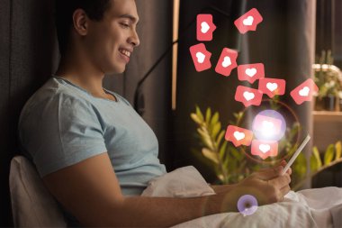 smiling mixed race man using digital tablet in bed near virtual hearts as likes illustration  clipart