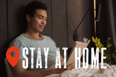 happy mixed race man reading book near stay at home lettering in bedroom  clipart