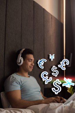 Kulaklıkla müzik dinleyen ve sanal para işaretlerinin yanında dizüstü bilgisayar kullanan iki ırklı bir adam.
