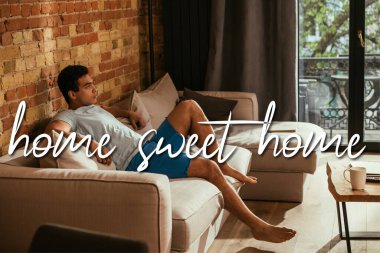 handsome mixed race man sitting on sofa in living room near home sweet home lettering  clipart