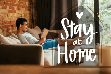 happy mixed race man reading book near stay at home lettering in living room  clipart