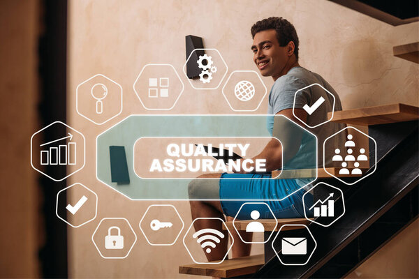 happy mixed race man using laptop on stairs near quality assurance lettering and virtual icons 