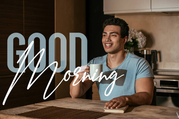 Счастливый смешанный человек, держащий чашку кофе рядом с книгой и добрым утром.
 