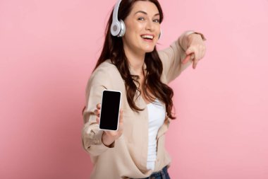 Kulaklıklı neşeli kadının seçici odağı pembe arka plandaki akıllı telefonu işaret ediyor.