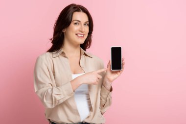 Gülümseyen kadın pembe arka planda akıllı telefonu işaret ediyor, vücut pozitifliği kavramı. 