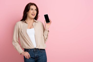 Çekici esmer kadın gülümserken pembe arka planda boş ekranlı akıllı telefon gösteriyor.
