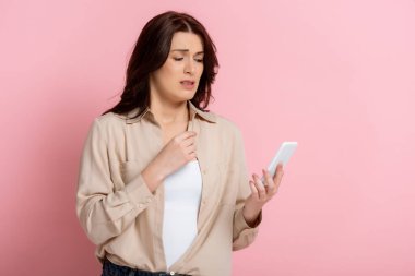 Worried woman holding smartphone on pink background, concept of body positive  clipart