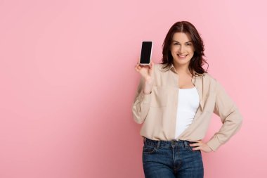 Güzel bir kadın kameraya gülümserken pembe arka planda boş ekranlı akıllı telefonu gösteriyor.