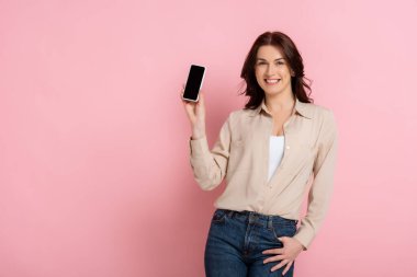 Güzel esmer kadın pembe arka planda akıllı telefonu tutarken kameraya gülümsüyor.