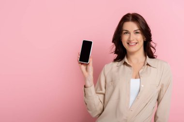 Çekici esmer kadın kameraya gülümserken pembe arka planda boş ekranlı akıllı telefon gösteriyor.
