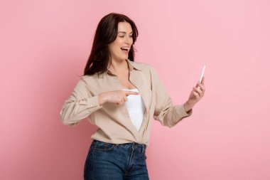 Positive woman pointing with finger at smartphone on pink background clipart
