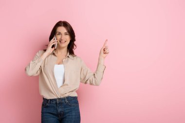 Güzel esmer kadın pembe arka planda akıllı telefonla konuşurken parmağıyla işaret ediyor.