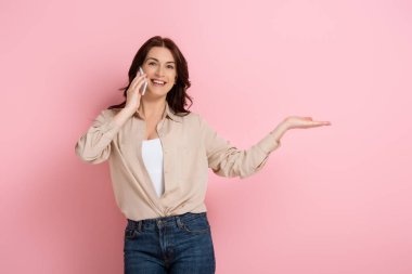 Beautiful smiling woman pointing with hand while talking on smartphone on pink background clipart