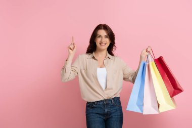 Pozitif esmer kadın pembe arka planda alışveriş torbalarını tutarken parmağıyla işaret ediyor.