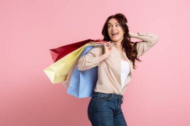 Elleri başının yakınında olan neşeli esmer kadın pembe arka planda renkli alışveriş torbaları tutuyor.
