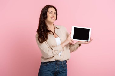 Güzel, gülümseyen bir kadın pembe arka planda boş ekran ile dijital tablet gösteriyor