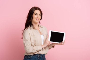 Esmer kadın, elinde pembe arka planda dijital tablet tutarken kameraya gülümsüyor