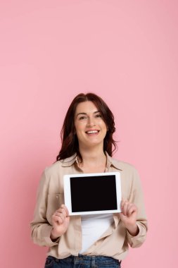 Güzel, gülümseyen bir kadın elinde pembe arka planda boş ekranlı dijital tablet tutuyor.