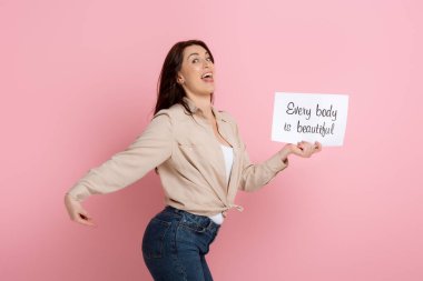 Happy woman pointing with finger on butt while holding card with every body is beautiful lettering on pink background