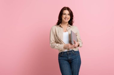 Çekici bir kadın pembe arka planda dizüstü bilgisayarını tutarken gülümsüyor.