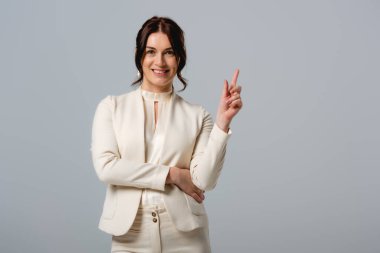 Beautiful businesswoman smiling at camera and pointing with finger isolated on grey  clipart