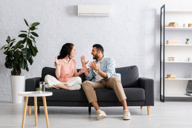 Positive couple smiling at each other while sitting on sofa under air conditioner at home  clipart