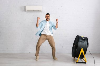Positive man showing yes gesture while standing near air conditioner and electric fan in living room clipart