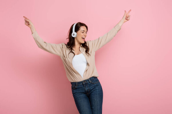 Cheerful woman pointing with fingers while listening music in headphones on pink background