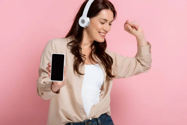 ヘッドフォンダンスの笑顔の女性とピンクの背景に空白の画面でスマートフォンを示す — ストック写真