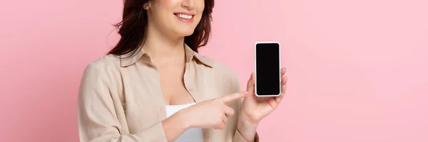 一个面带微笑的女人用手指指指用粉色隔离的智能手机的全景 — 图库照片