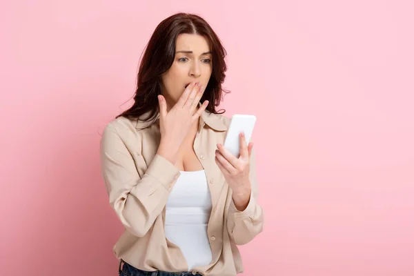 ショックを受けたブルネット女性とともに手近く口の中でスマートフォンを使用してピンクの背景 概念の体陽性 — ストック写真