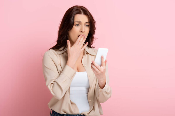 Shocked brunette woman with hand near mouth using smartphone on pink background, concept of body positive 