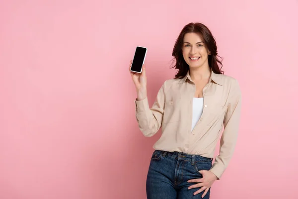 漂亮的黑发女人一边拿着粉色背景的智能手机一边对着相机微笑 — 图库照片