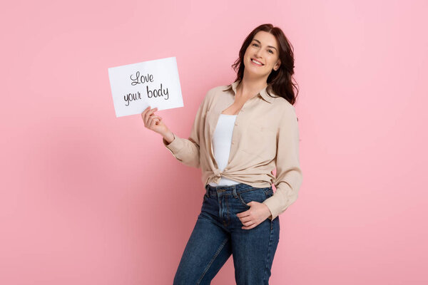 Smiling woman holding card with love your body lettering on pink background, concept of body positive
