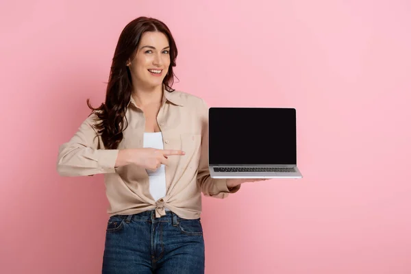 微笑的女人用手指指指笔记本电脑 粉红背景上的空白屏幕 — 图库照片