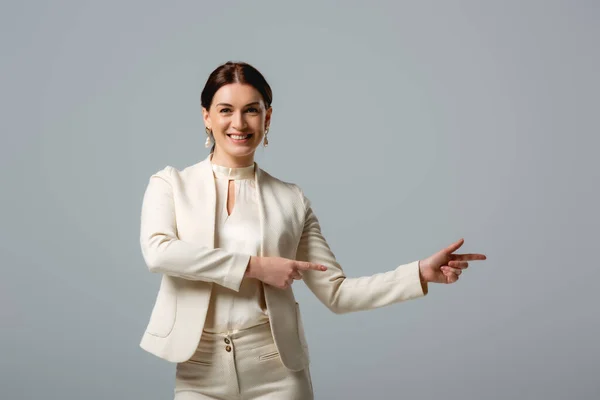 Smiling woman in formal wear pointing with fingers isolated on grey