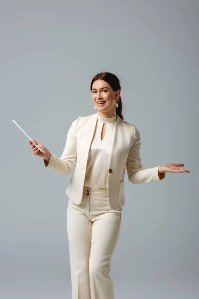 Attractive woman in formal wear smiling at camera while holding digital tablet isolated on grey, concept of body positive