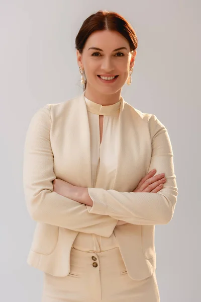 Positive brunette woman in formal wear with crossed arms smiling at camera isolated on grey