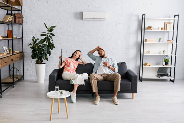 Young woman holding fan near boyfriend with glass of water while suffering from heat at home 