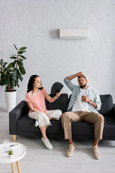 Utmattad Man Håller Glas Vatten Nära Flickvän Med Fläkt Soffan — Stockfoto