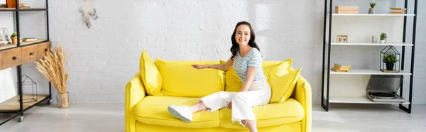 自宅で黄色のソファに座っている間にカメラで微笑む若い女性のパノラマショット — ストック写真