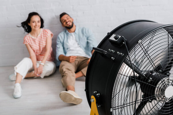 Selective focus of electric fan near smiling couple sitting on floor at home 