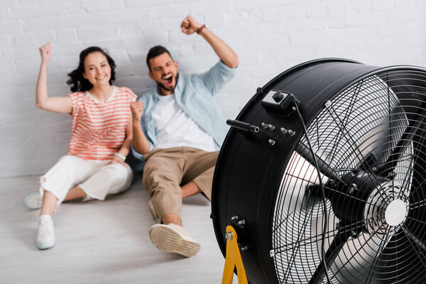 Selective focus of cheerful couple showing yeah gesture while sitting near electric fan on floor 