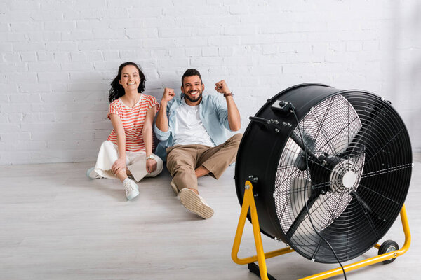 Young couple smiling at camera and showing yeah gesture while sitting on floor near electric fan 