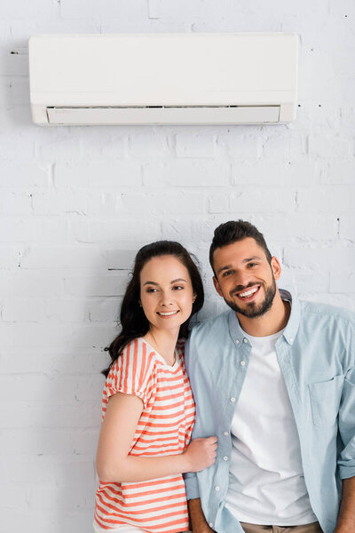 Handsome man smiling at camera near girlfriend and air conditioner on wall 