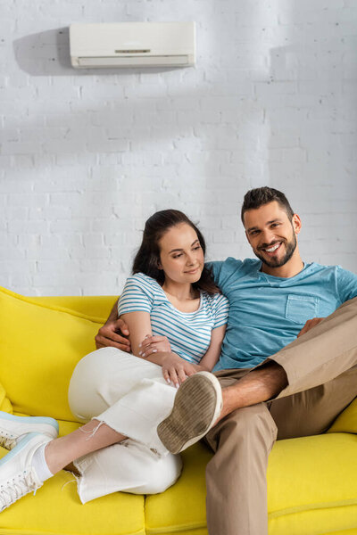 Handsome man smiling at camera while hugging attractive girlfriend on couch in living room