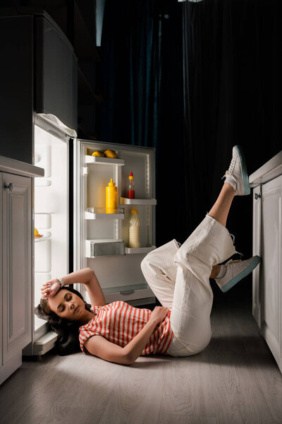 Beautiful woman lying on floor near open fridge in kitchen at night