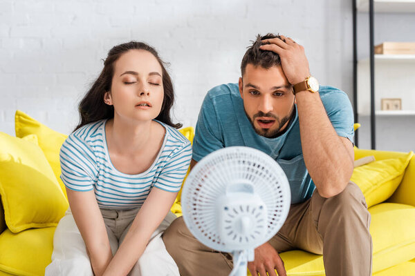 Selective focus of young couple sitting near electric fan in living room