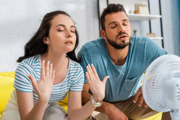 Selective focus of young couple sitting near electric fan in living room