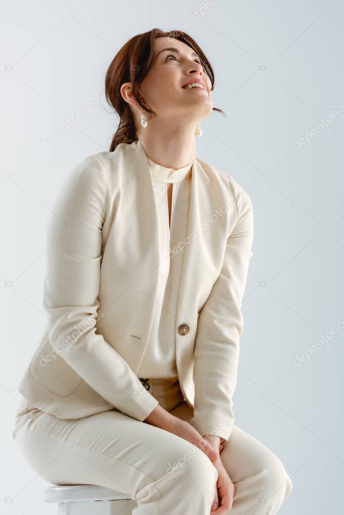 Positive businesswoman looking up while sitting on chair isolated on grey 
